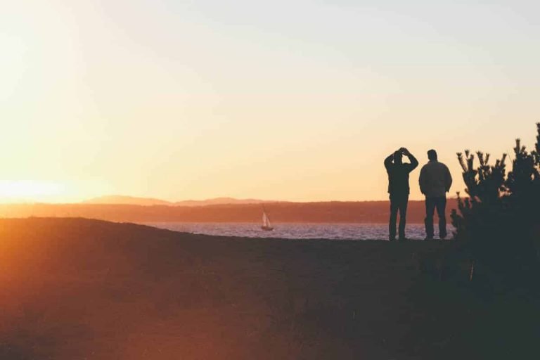 Deux personnes debout sur une colline au coucher du soleil, face à l'eau avec un voilier au loin. Ambiance calme et chaleureuse.