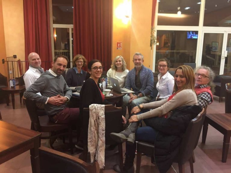 Dix adultes souriants autour d'une table, avec ordinateurs, en formation Gestalt-thérapie dans une salle chaleureuse.