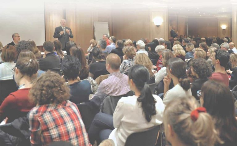 Un grand groupe est assis de face dans une salle de conférence bien éclairée, écoutant attentivement un conférencier de l'École Humaniste de Gestalt qui se tient près d'un tableau blanc, tandis que des rangées de participants s'intéressent aux concepts de la Gestalt-thérapie.