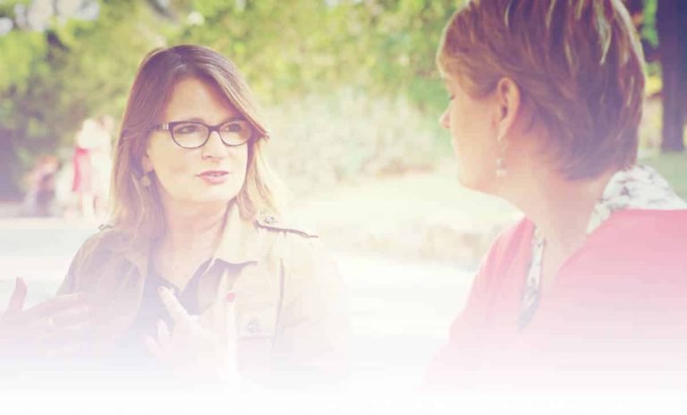 Deux femmes sont assises à l'extérieur et discutent ; l'une d'elles, qui porte des lunettes, fait des gestes expressifs tandis que l'autre l'écoute attentivement, reflétant un moment de connexion que l'on trouve dans la formation Gestalt Thérapie. L'arrière-plan flou et ensoleillé et le premier plan doux créent un effet aérien.