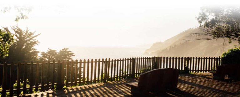 Une clôture en bois et des bancs surplombent une côte pittoresque et brumeuse, avec de grands arbres et des collines en arrière-plan, baignés par la lumière chaude du soleil - une scène tranquille qui invite à la réflexion, tout comme la perspective holistique adoptée dans la pratique de la Gestalt.