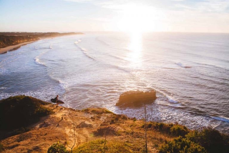Un surfeur solitaire se tient sur une falaise surplombant l'océan au coucher du soleil, s'imprégnant de la lumière dorée - un moment de pleine conscience qui rappelle la Gestalt, où l'on devient pleinement présent et connecté à ce qui nous entoure.