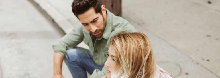 Un homme accroupi sur un trottoir de la ville regarde avec inquiétude une femme à côté de lui qui semble bouleversée - une interaction qui fait écho à l'écoute compatissante enseignée dans la formation à la thérapie gestaltiste. Tous deux sont habillés de façon décontractée et sont engagés dans une conversation sérieuse.