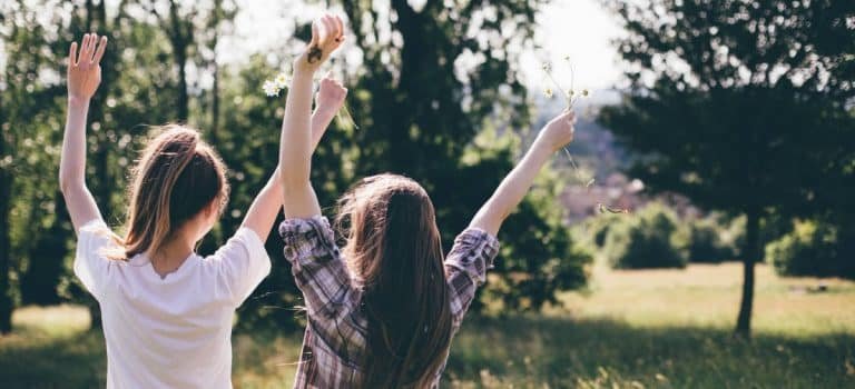 Deux jeunes filles se tiennent en plein air dans un champ herbeux, les bras levés tenant des fleurs sauvages - un moment qui fait écho à l'esprit de la Gestalt thérapie. La lumière du soleil coule tandis que les arbres encadrent l'arrière-plan, capturant un sentiment de connexion et de présence attentive.