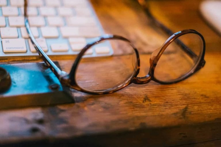 Une paire de lunettes rondes et brunes repose sur un bureau en bois à côté d'une partie d'un clavier d'ordinateur blanc et d'un support d'angle en métal, évoquant l'ambiance réfléchie de la Formation gestalt thérapie.