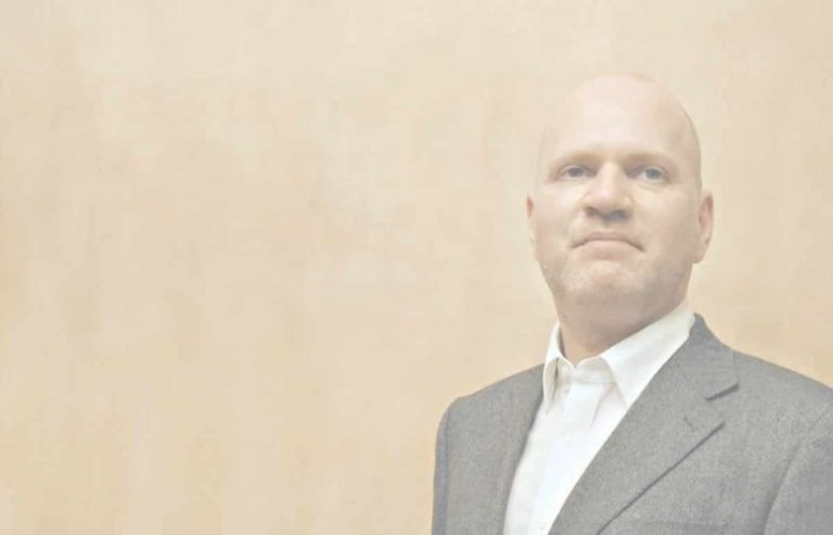 Un homme vêtu d'une veste de costume grise et d'une chemise blanche se tient devant un fond beige clair uni, le regard légèrement tourné vers la gauche. L'image évoque une présence calme, idéale pour promouvoir une formation gestalt thérapie.