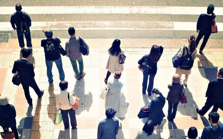 Un groupe de personnes vues de haut, debout sur un trottoir, attendant à un passage pour piétons. La scène évoque la connexion et la prise de conscience, valeurs fondamentales de la Gestalt-thérapie. La lumière du soleil projette de longues ombres derrière eux sur l'arrière-plan visible de la rue.