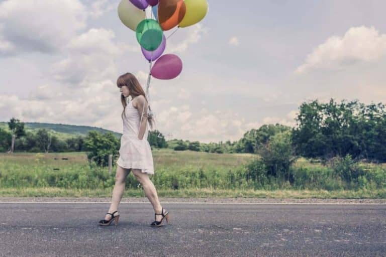 Une jeune femme vêtue d'une robe blanche marche le long d'une route déserte en tenant des ballons colorés - une scène paisible qui fait écho à la découverte de soi dans le cadre de la Gestalt thérapie, avec des champs verts, des arbres et un ciel partiellement nuageux en arrière-plan.