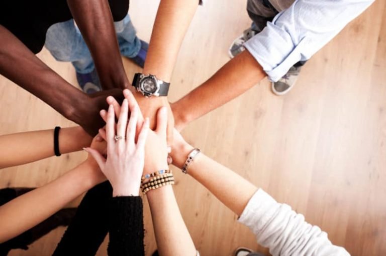 Un groupe de personnes diverses se tenant en cercle place leurs mains ensemble au centre, symbolisant l'unité et le travail d'équipe, reflétant l'esprit de la Gestalt à l'École Humaniste de Gestalt, sur un fond de plancher en bois.
