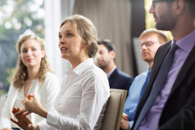 Une femme en chemise blanche prend la parole, assise au milieu d'un petit groupe qui l'écoute attentivement, dans un cadre professionnel ou un séminaire axé sur la Gestalt-thérapie.