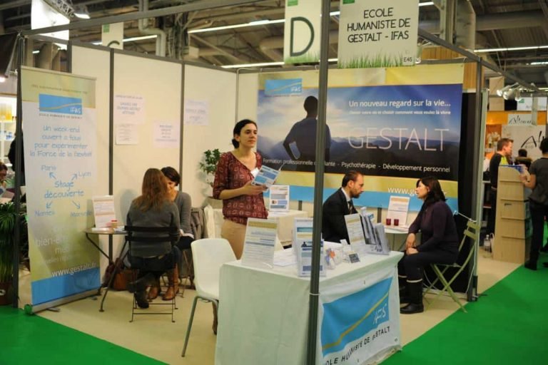 Une femme tient des brochures sur un stand de l'École Humaniste de Gestalt lors d'une exposition, où plusieurs personnes discutent de la Gestalt thérapie et de la formation gestalt praticien, entourées de bannières et de documents sur la psychothérapie et le développement personnel.