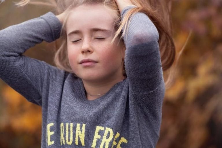 A young girl with closed eyes and hands in her hair stands outdoors, wearing a grey sweater with yellow text that reads BE RUN FREE. The blurred autumn background evokes a sense of presence akin to Gestalt thérapie.