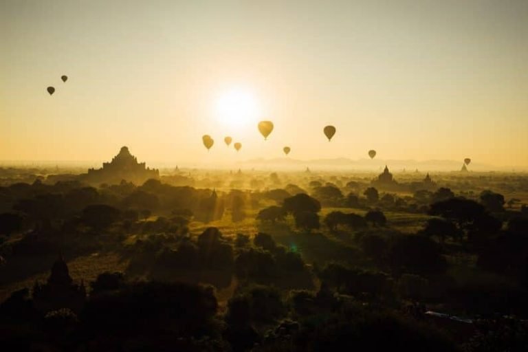 Hot air balloons float over ancient temples and trees at sunrise, much like the transformative journey of a formation gestalt praticien—casting long shadows across a vast landscape bathed in golden light.