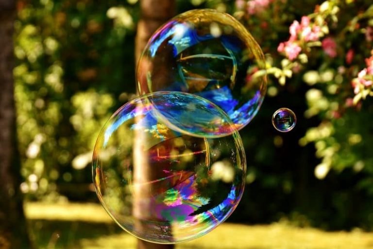 Two large, colorful soap bubbles float in the air with a small bubble nearby. The softly blurred background of green foliage, tree trunk, and pink flowers evokes the gentle perspective often explored in Gestalt thérapie.