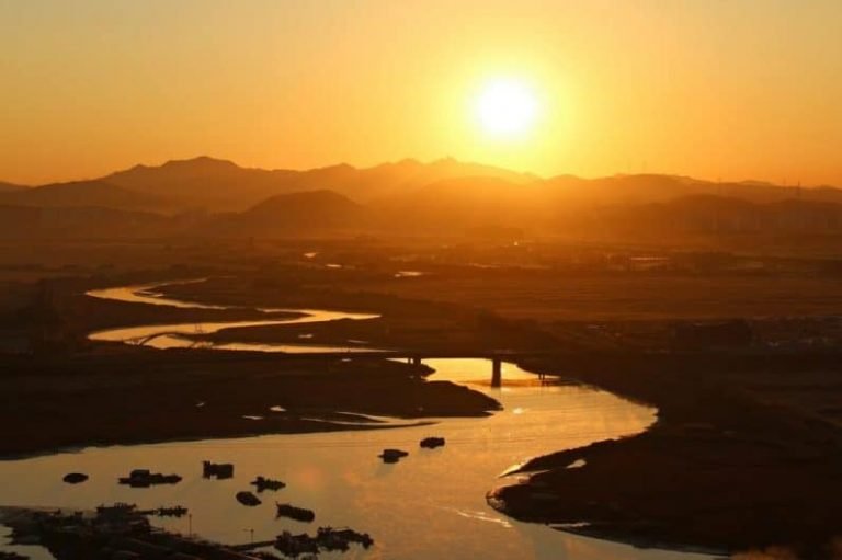 Une rivière sinueuse reflète la lumière dorée du soleil couchant avec, en arrière-plan, des silhouettes de montagnes et un ciel brumeux, évoquant l'atmosphère tranquille souvent explorée dans la Formation gestalt thérapie. Le paysage est baigné de tons chauds et orangés.