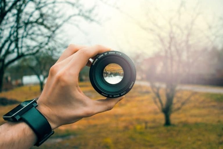 A hand wearing a black smartwatch holds a camera lens, with trees and grass visible through the lens. The blurred background highlights the focus—much like Gestalt thérapie, emphasizing clarity and perspective in understanding experiences.