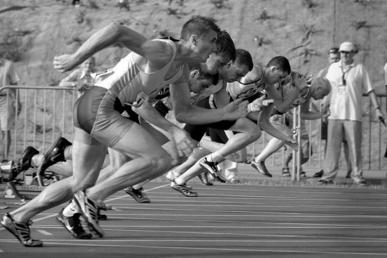 Six male athletes in running gear sprint off the starting blocks in a track race, captured in mid-motion. A few officials and spectators, reflecting Gestalt awareness of the present moment, stand in the background near a barrier. The image is in black and white.
