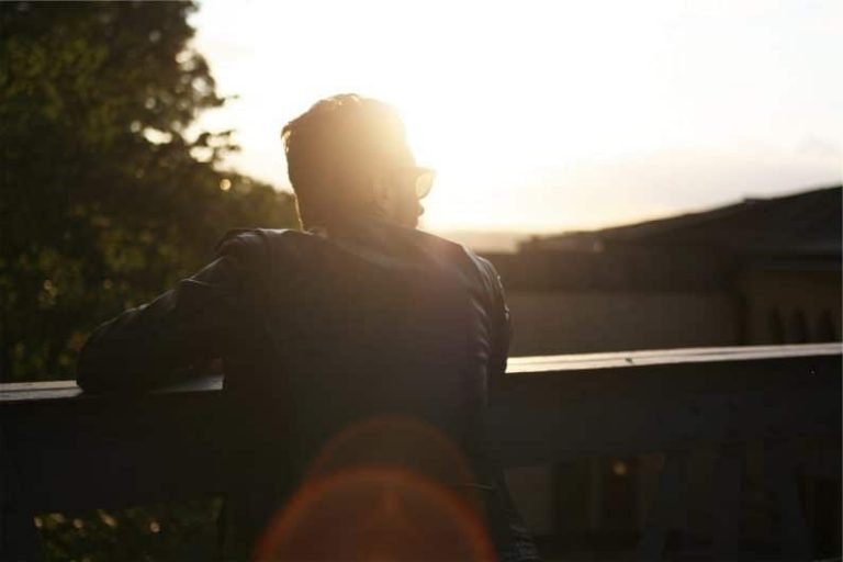 A person wearing sunglasses and a jacket leans on a wooden railing, looking towards the bright setting sun, reflecting on their journey in formation gestalt thérapie, with trees and buildings in the background.