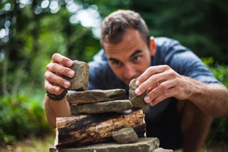 A man crouches outdoors, carefully stacking flat stones and pieces of wood, his focus echoing the mindful balance encouraged in Gestalt thérapie, with a green, blurred forest background.