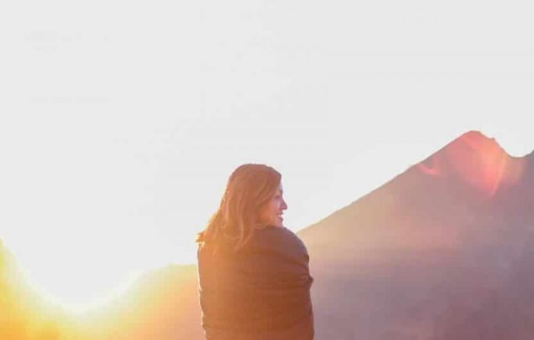 Une personne enveloppée dans une couverture se tient à l'extérieur au lever ou au coucher du soleil, avec des montagnes en arrière-plan et la lumière du soleil illuminant la scène - embrassant un nouveau jour, tout comme on commence son voyage avec l'École Humaniste de Gestalt.