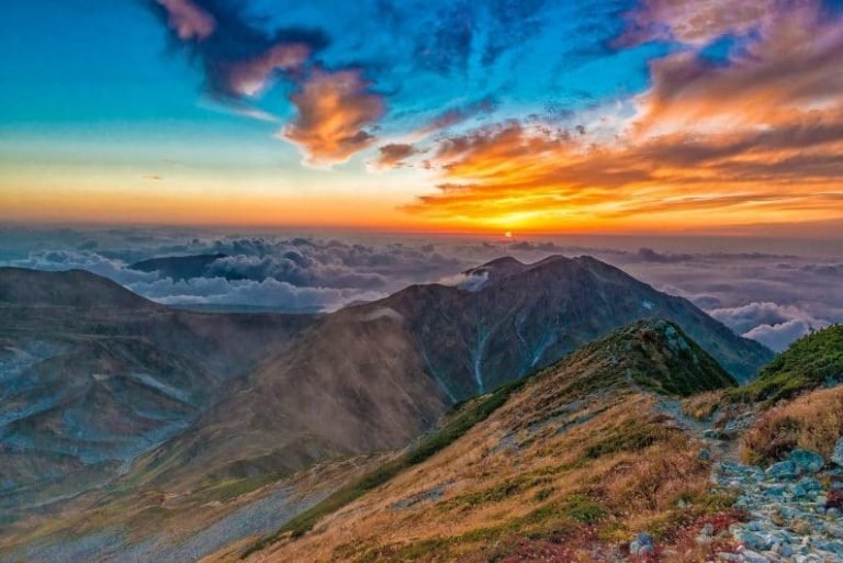 A vibrant sunrise over mountain peaks, with colorful clouds and sun rays illuminating the sky. Mist fills the valleys below, and grassy slopes with rocky paths evoke a sense of discovery—much like a journey in Gestalt thérapie.