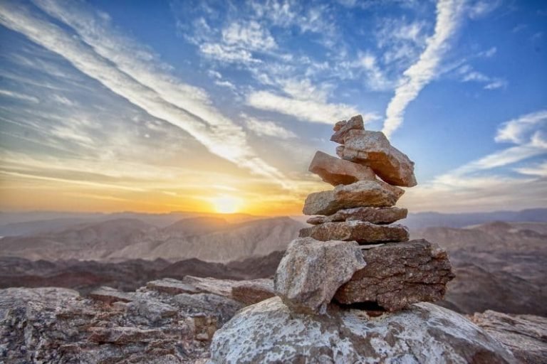 Une petite pile de rochers se tient en équilibre sur le sommet d'une montagne, avec en arrière-plan un lever ou un coucher de soleil spectaculaire, évoquant les principes de la Gestalt sous un ciel coloré parsemé de nuages vaporeux et de crêtes montagneuses lointaines.
