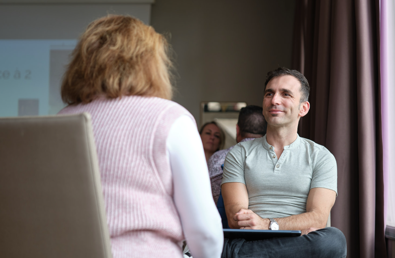 A man and woman sit facing each other indoors, engaged in conversation. The man, prenant le rôle thérapeute, is smiling and listening attentively, while other people are visible in the background.
