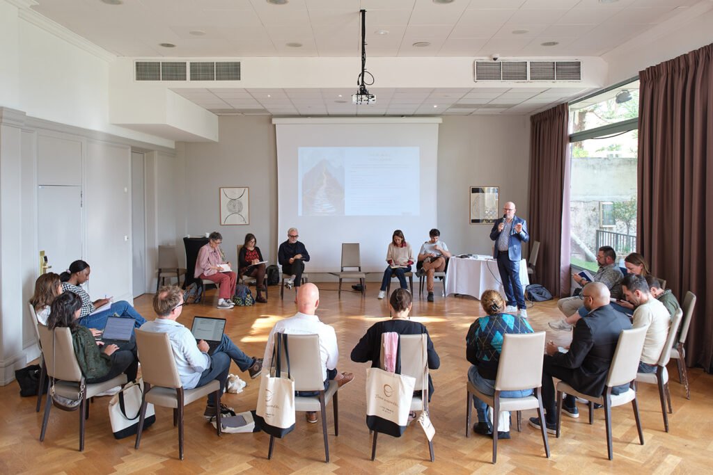 A group of people sits in a circle in a bright conference room, listening to a speaker discuss dépendance affective near a screen displaying a presentation. Some attendees use laptops and notepads, and large windows let in natural light.