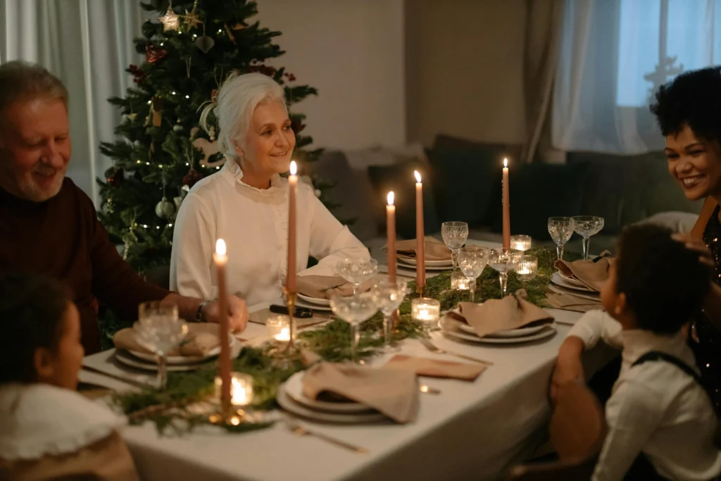 A family sits around a candle-lit dinner table decorated with greenery, celebrating together near a Christmas tree. Smiling and engaged in conversation, they create a warm and festive atmosphere perfect for holiday fêtes.