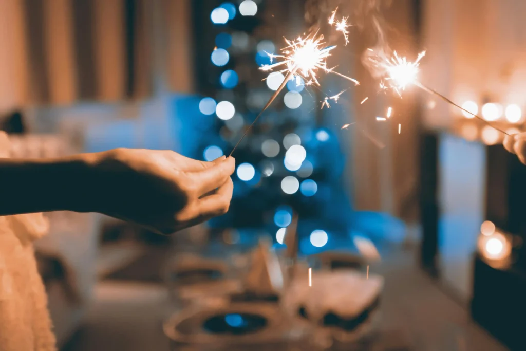 Two hands hold lit sparklers in a warmly lit room, with a blurred, decorated Christmas tree and festive lights in the background, evoking both the joy of celebration and the tenderness of moments tinged by dépendance affective.