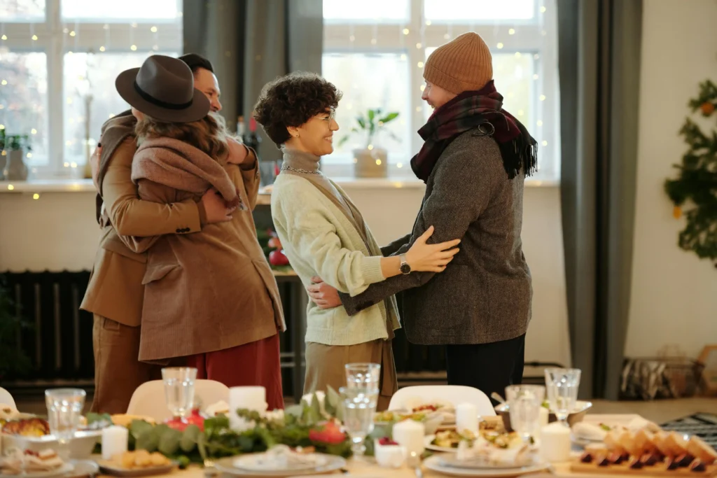 Four people greet each other warmly indoors, two pairs hugging and smiling, in a festively decorated room with a table set for a meal in the foreground. Holiday lights and greenery add to the cozy atmosphere of shared joy and dépendance affective.