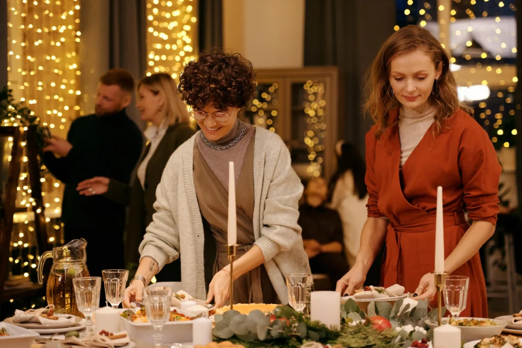 Two women set a festive dinner table with candles, glasses, and plates of food, while fêtes come alive in the background amid people and warm string lights, creating a cozy, celebratory atmosphere.
