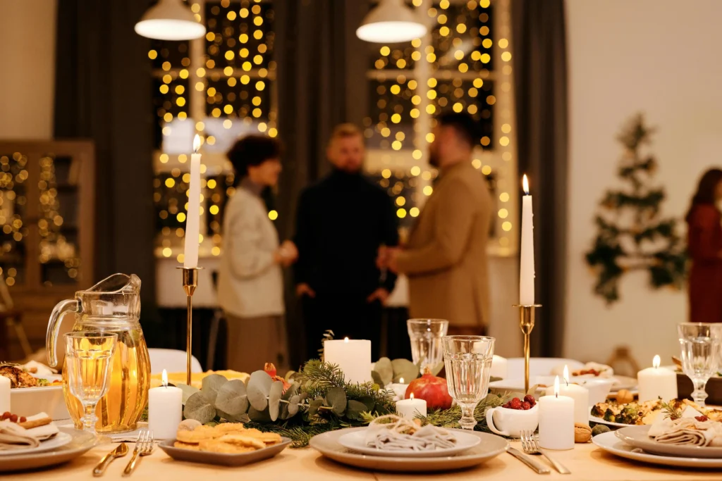 A festive dinner table with candles, food, and drinks is set in the foreground, while three people—bound by amitié and perhaps a touch of dépendance affective—stand talking near twinkling lights and a decorated tree in the softly lit background.