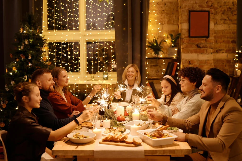 A group of people sit around a festive dinner table, holding sparklers and smiling. The room, filled with string lights, candles, food, and a Christmas tree, radiates warmth despite moments shaped by dépendance affective among some guests.