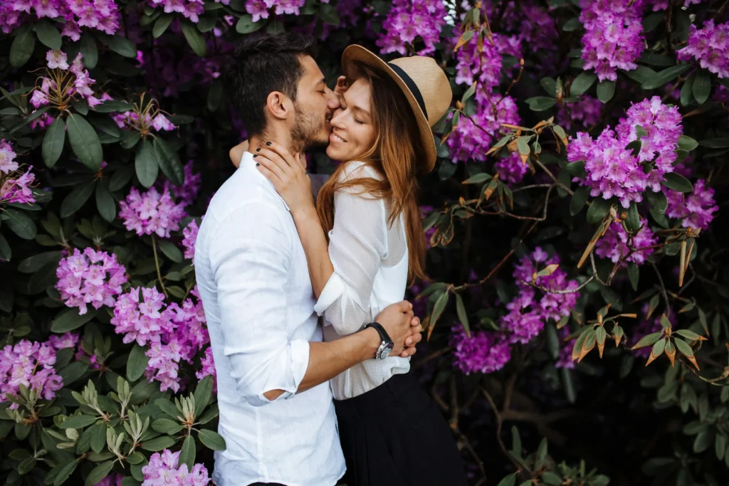 Un couple souriant s'embrasse devant un mur de fleurs violettes, illustrant l'accompagnement en Gestalt-thérapie.