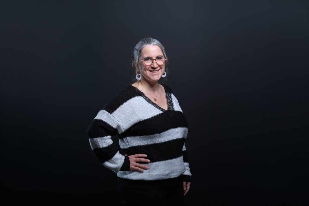 Femme souriante aux cheveux gris et lunettes, pull rayé noir-blanc, debout main sur la hanche devant fond uni sombre.