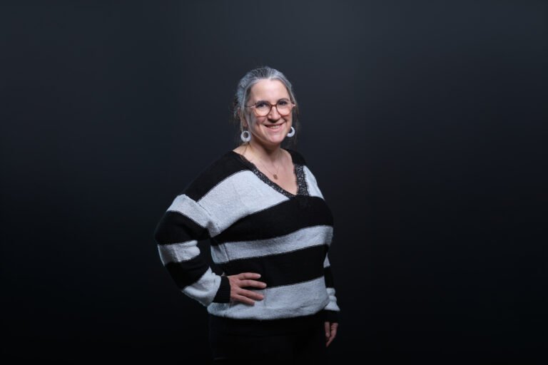 Femme souriante aux cheveux gris et lunettes, pull rayé noir-blanc, debout main sur la hanche devant fond uni sombre.