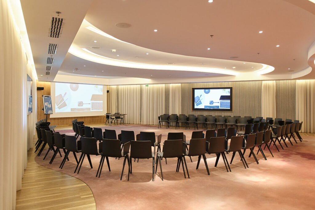 Salle de conférence moderne, sièges noirs circulaires, deux écrans, sol bois, rideaux neutres et éclairage doux.