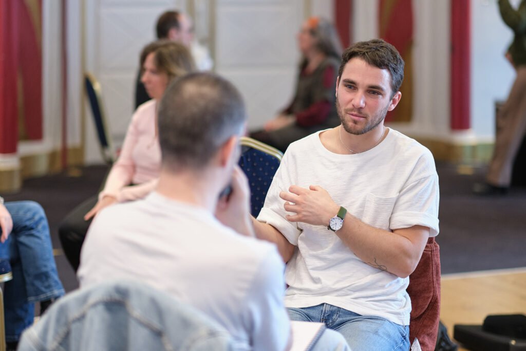 Deux hommes échangent face à face lors d'un atelier de Gestalt-thérapie, d'autres participants discutent en arrière-plan.