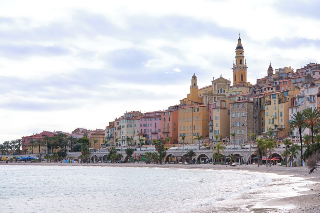 Des bâtiments colorés sur une colline en bord de mer, une plage de sable, une mer calme, des palmiers et un clocher d’église décoré.