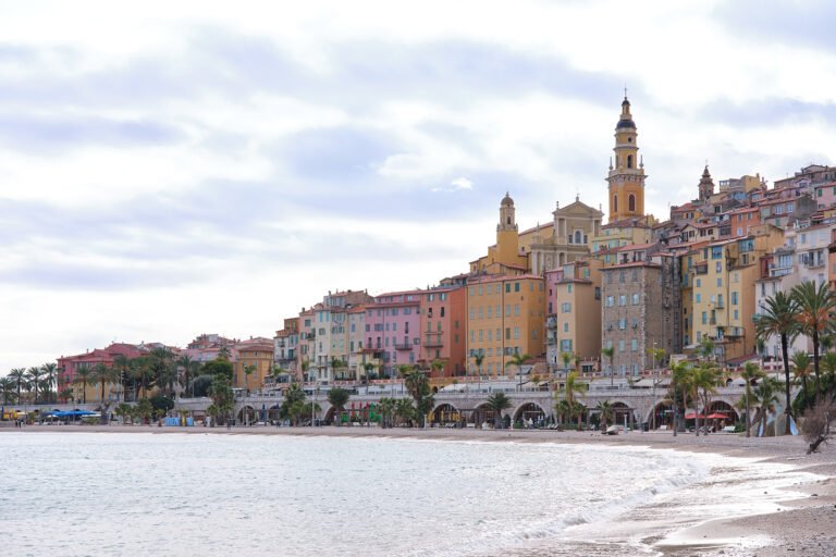 Des bâtiments colorés sur une colline en bord de mer, une plage de sable, une mer calme, des palmiers et un clocher d’église décoré.