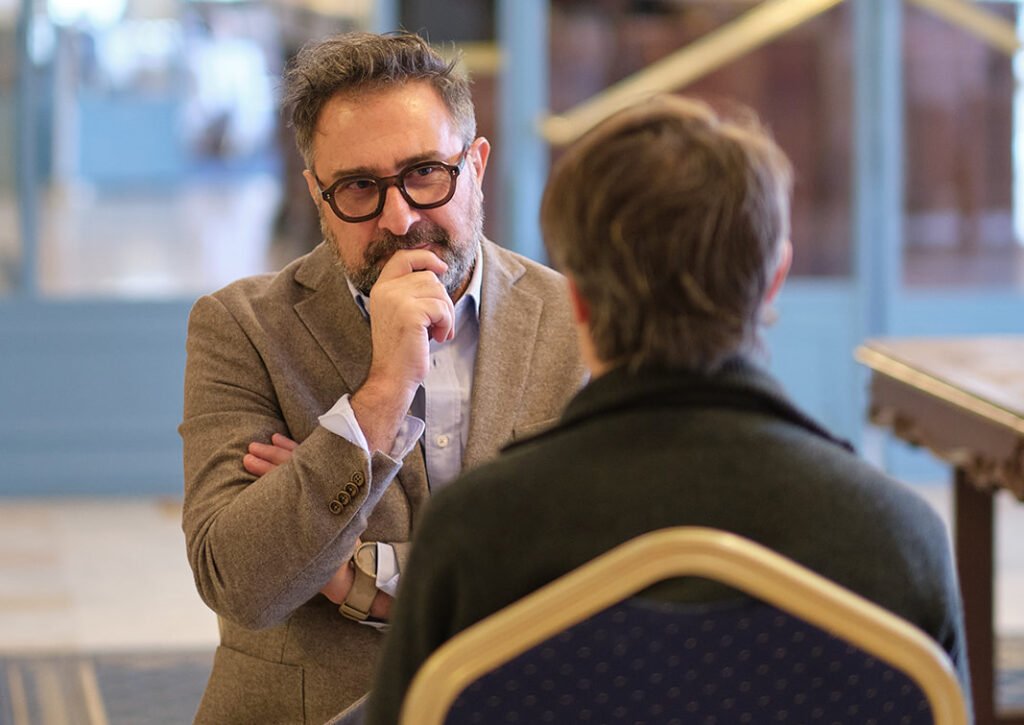 Deux hommes discutent dans une pièce élégante et lumineuse ; l’un, barbu et portant des lunettes, se touche le visage. Thérapie Gestalt.
