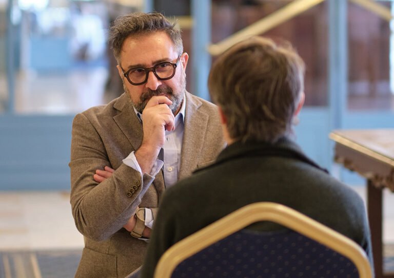 Deux hommes discutent dans une pièce élégante et lumineuse ; l’un, barbu et portant des lunettes, se touche le visage. Thérapie Gestalt.