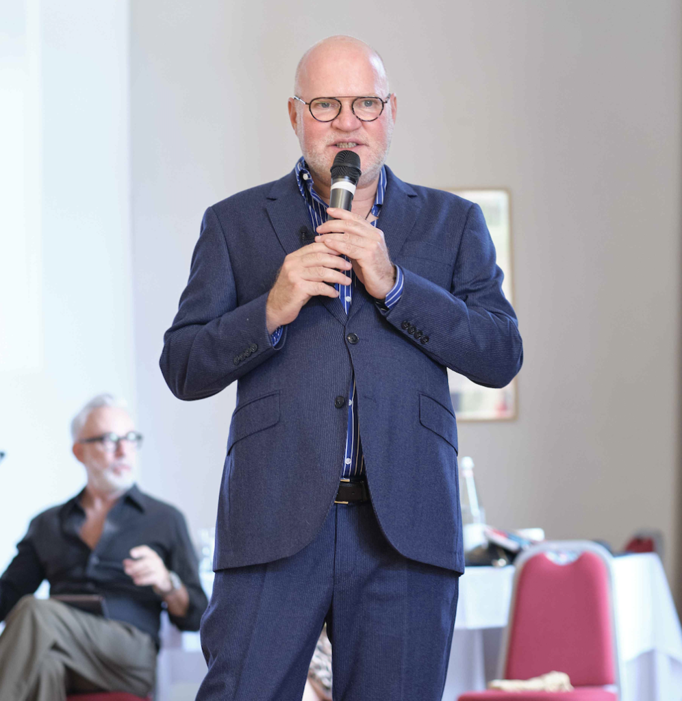 Un homme chauve, vêtu d'un costume bleu et portant des lunettes, parle dans un micro, debout devant des chaises rouges et une personne assise.