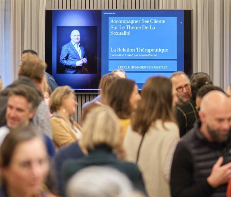 Des participants discutent lors d’un événement organisé en salle ; à l’arrière-plan, un écran montre un homme en costume ainsi qu’un texte en français sur la relation thérapeutique et une formation à la sexualité.