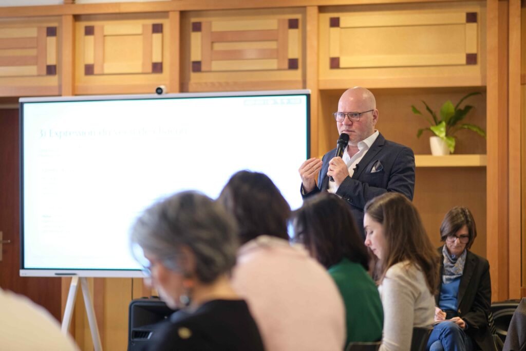 Un homme en blazer donnant une conférence sur la sexualité, micro à la main, devant un écran et un public participant à une formation en Gestalt.
