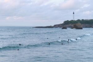 Des surfeurs en combinaison glissant sur les vagues de l'océan, avec un phare côtier en arrière-plan.