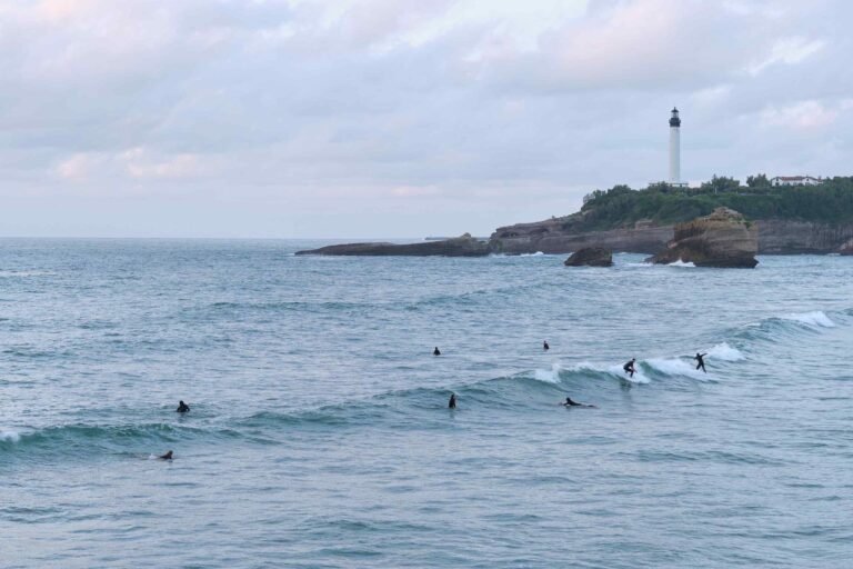 Des surfeurs en combinaison glissant sur les vagues de l'océan, avec un phare côtier en arrière-plan.
