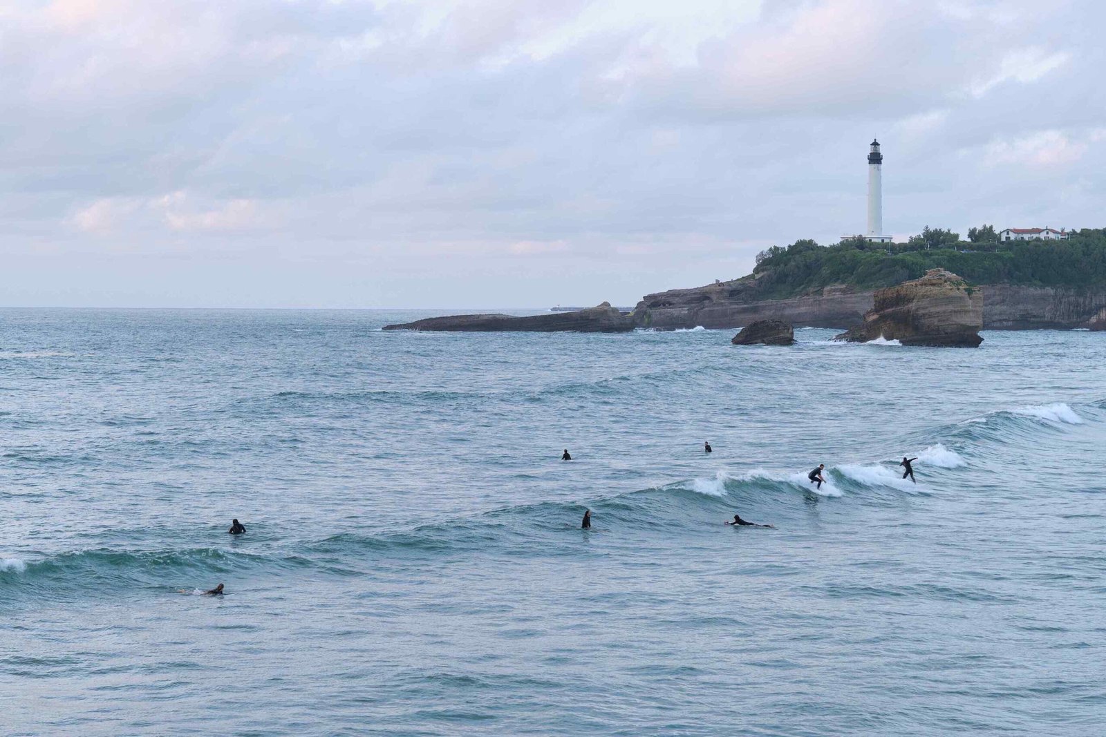 Des surfeurs en combinaison glissant sur les vagues de l'océan, avec un phare côtier en arrière-plan.